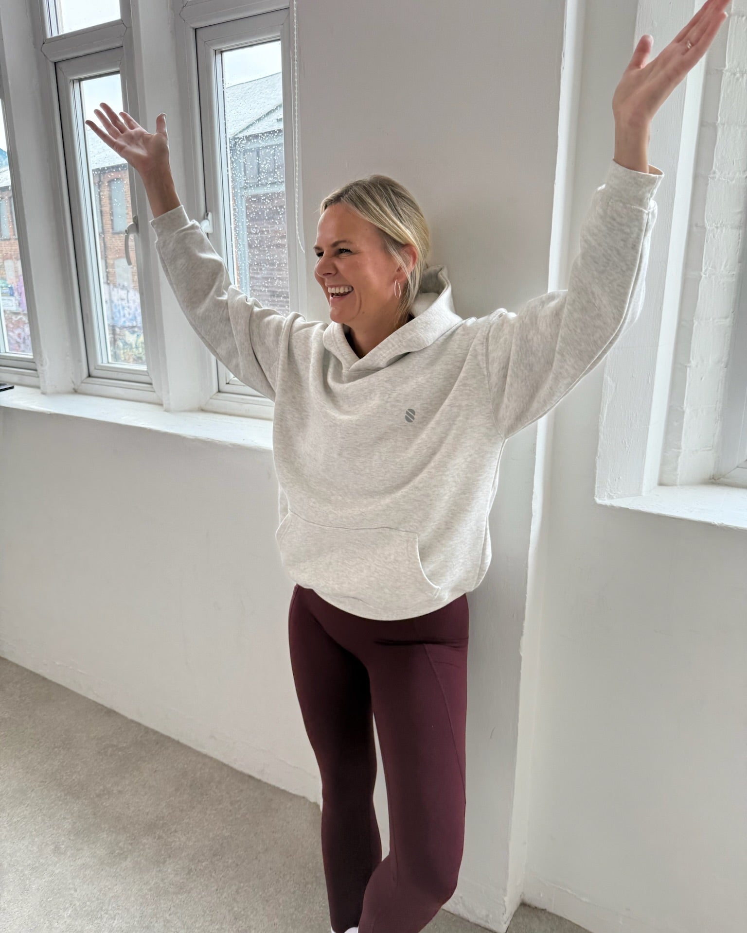 Person wearing a light gray hoodie and dark cherry leggings standing in a room with large windows.