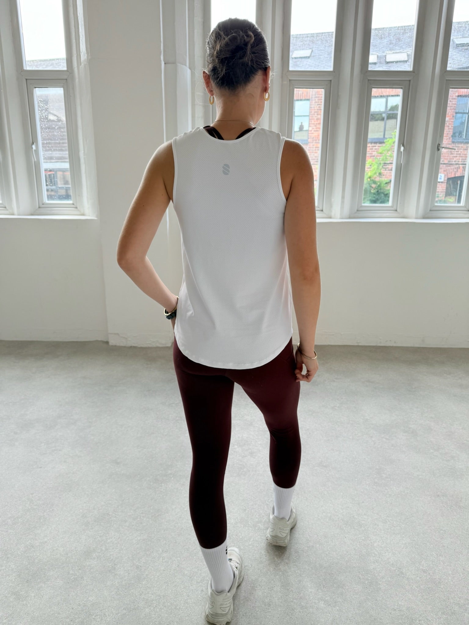 Person wearing a white tank top and maroon leggings standing in a room with large windows.