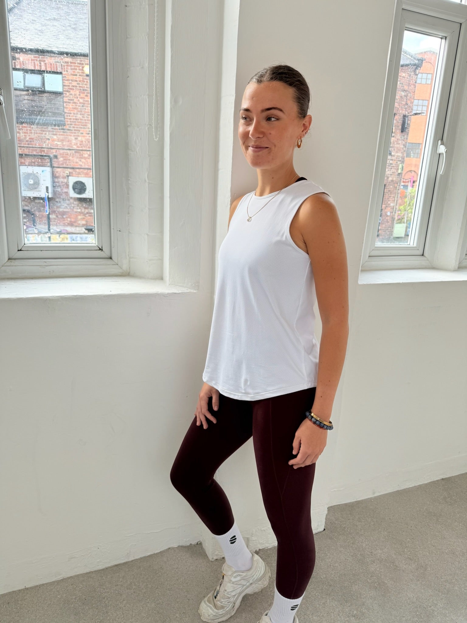 Woman in a white sleeveless top and dark leggings standing in a room with large windows.