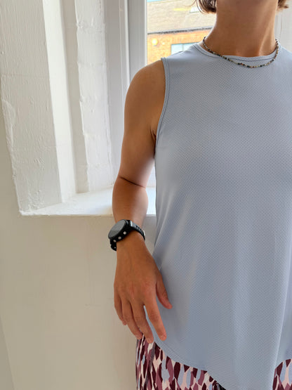 Person wearing a light blue sleeveless top and patterned pants, standing indoors with a window in the background.