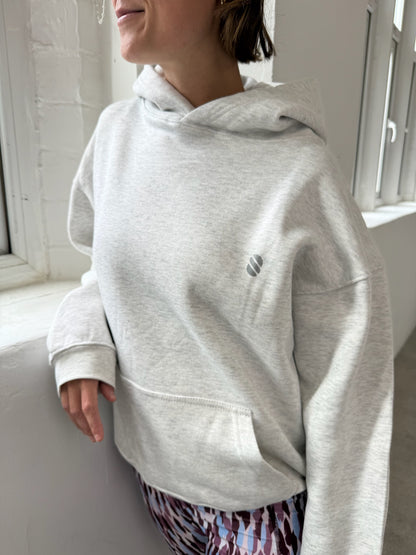 Person wearing a gray hoodie with a logo, standing indoors near a window.