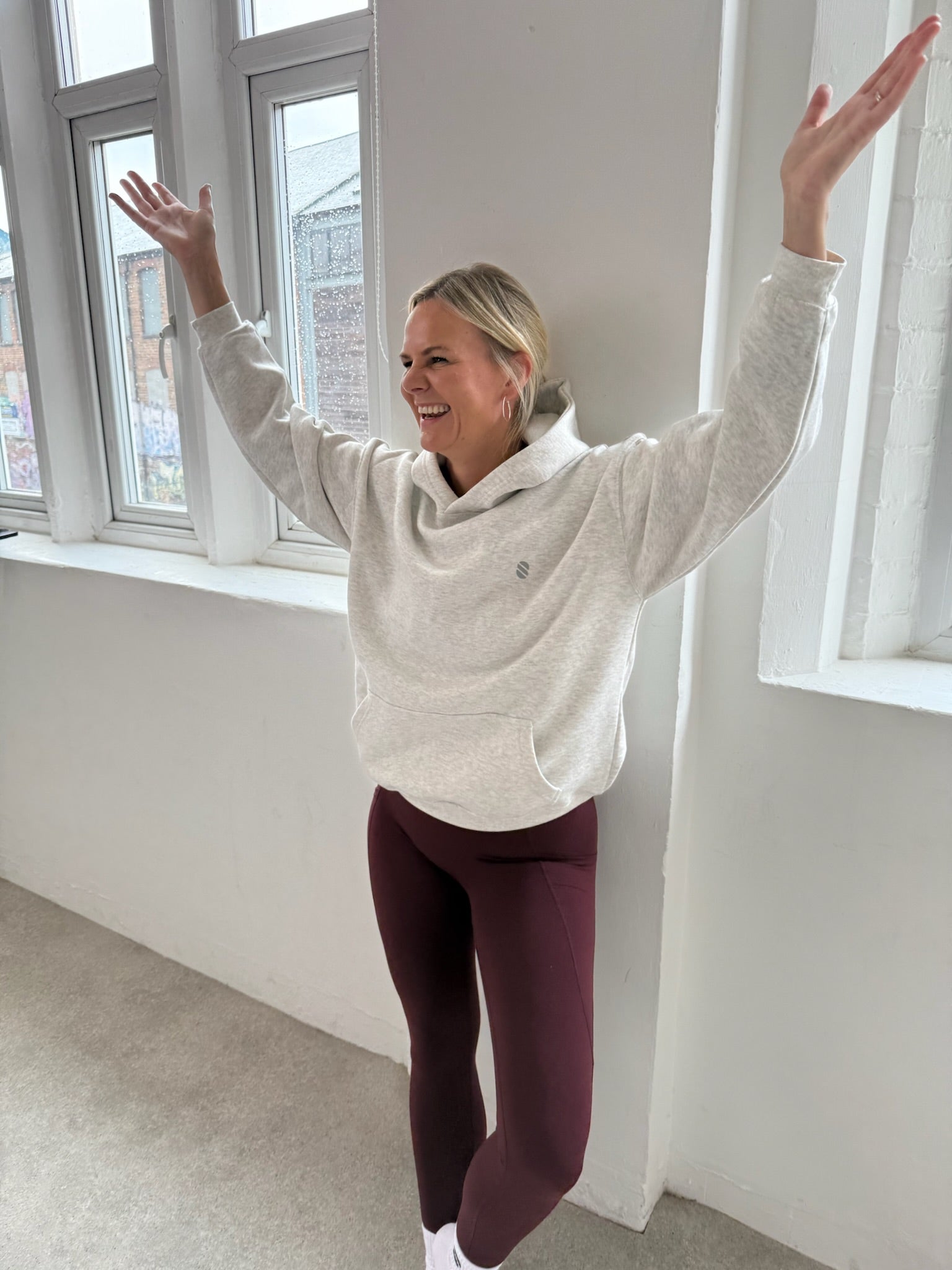 Person wearing a light gray hoodie and dark cherry leggings standing in a room with large windows.