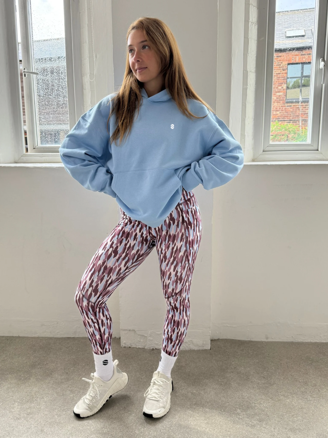 Person wearing a blue sweatshirt and patterned leggings standing in a room with large windows.
