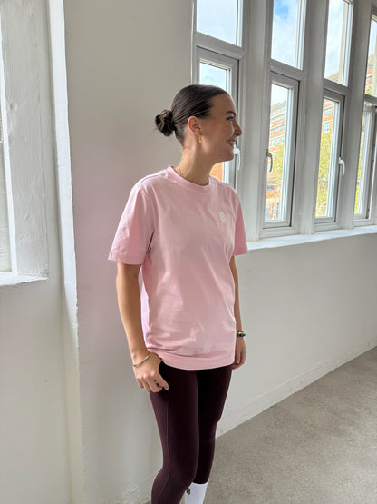 Person wearing a pink t-shirt and dark leggings standing in a room with large windows.