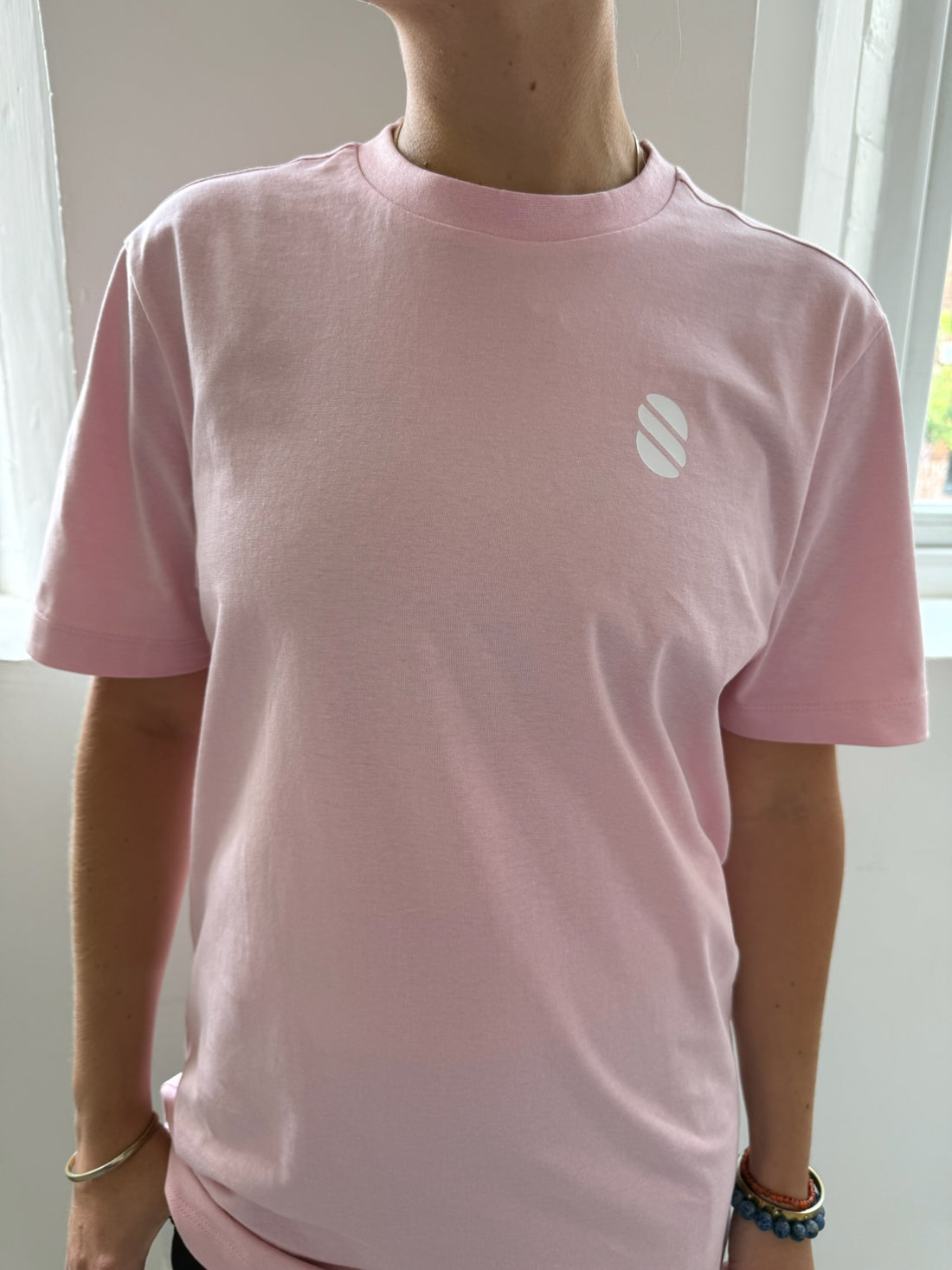 Person wearing a pink t-shirt with a white logo in front of a window.