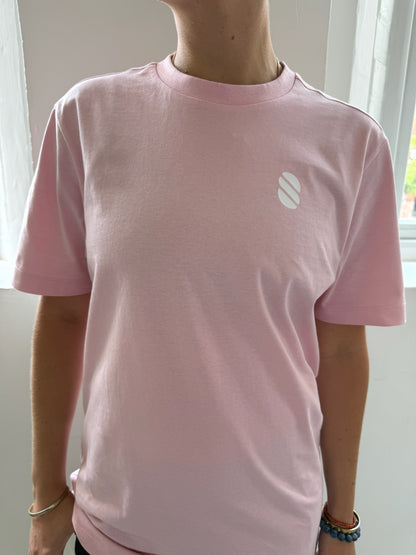 Person wearing a pink t-shirt with a white logo in front of a window.