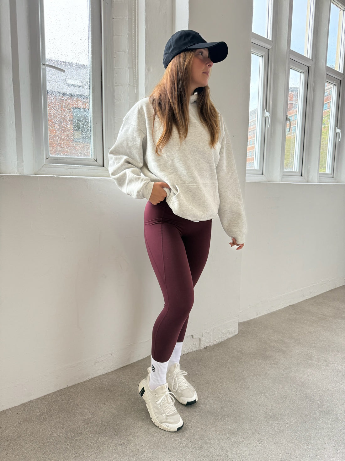Person wearing a white sweatshirt, maroon leggings, and white sneakers in a room with large windows.
