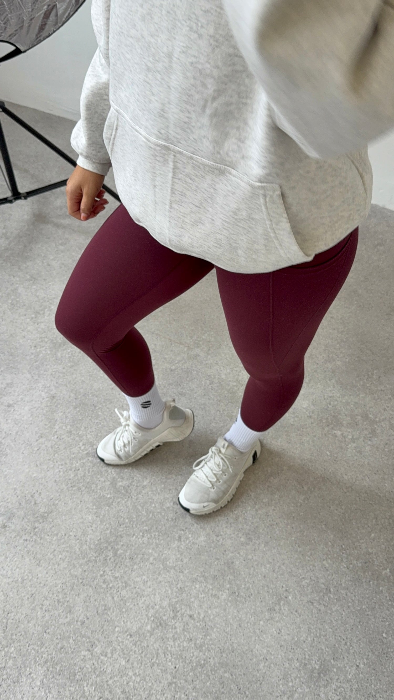 Person wearing maroon leggings, white sneakers, and a light gray hoodie on a neutral background