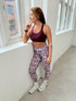 Woman in dark cherry sports bra and patterned leggings standing by a window in a bright room.